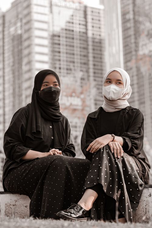 Free Two women wearing hijabs and masks sitting outdoors in a cityscape. Stock Photo