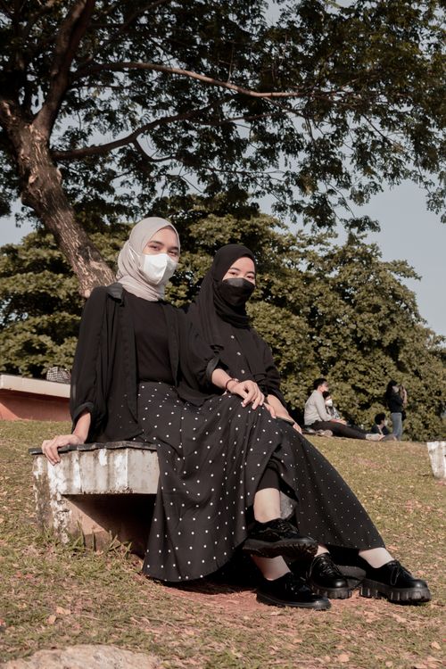 Free Two women in hijabs and face masks sit on a park bench outdoors. Stock Photo