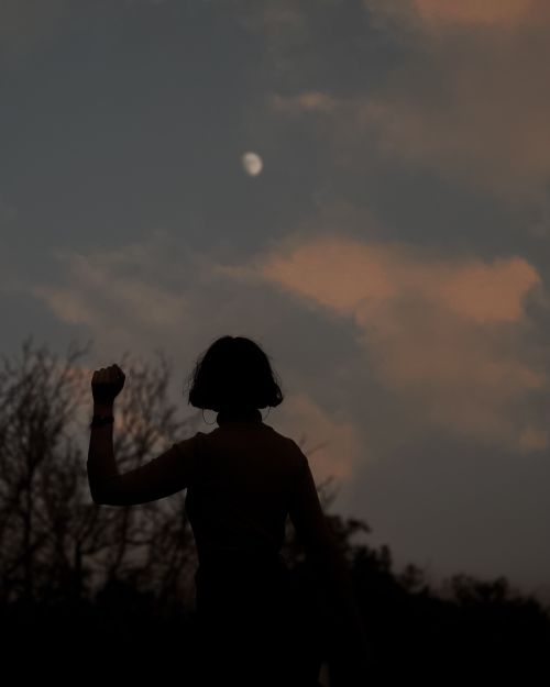 Free Silhouette of a person with a raised fist against a moonlit sky with scattered clouds. Stock Photo