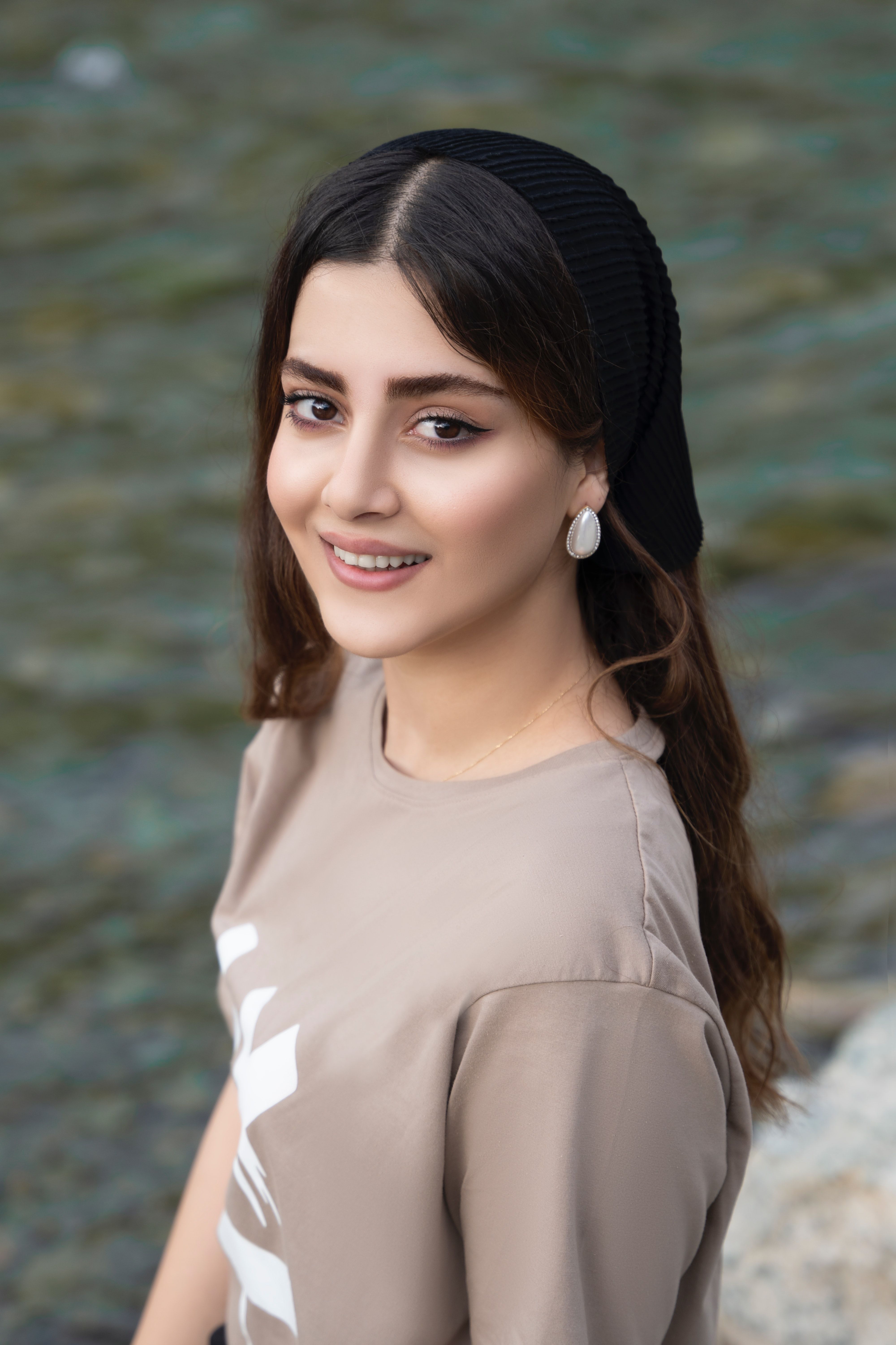 A smiling woman with dark hair poses outdoors by a river, wearing a headscarf and earrings.