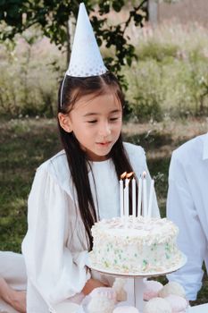 A young girl in a party hat blows candles on a birthday cake during a garden celebration.