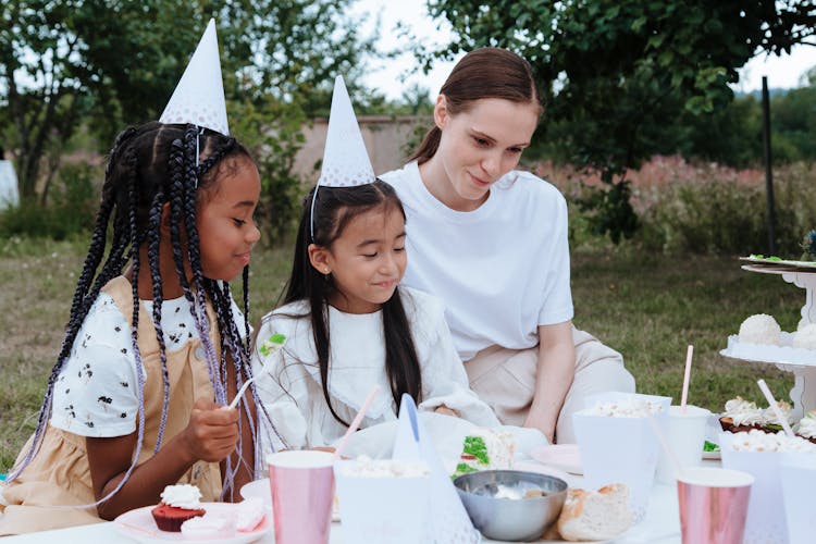 Woman And Two Girls In Party Hats Sitting At Table In Garden