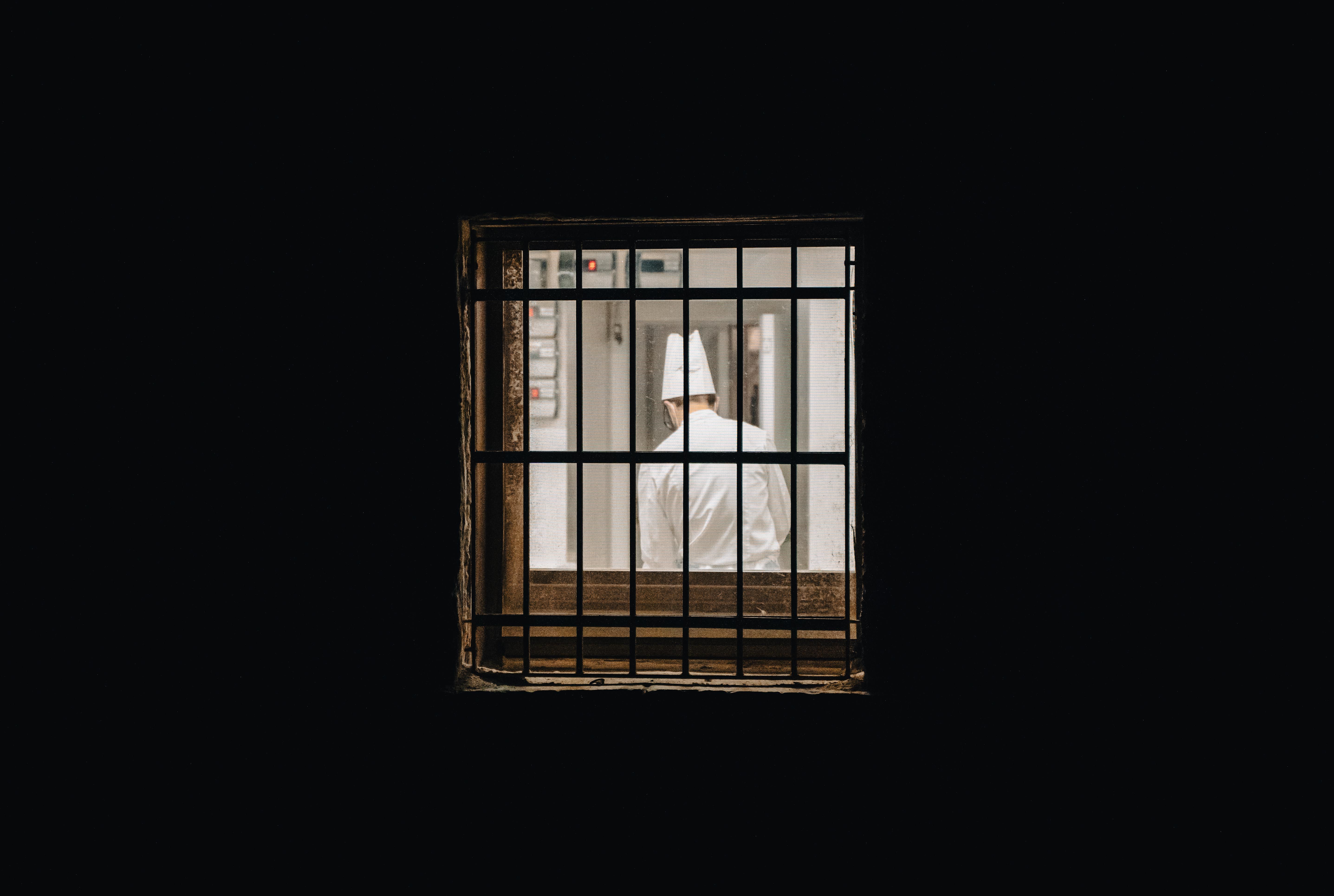A chef in a white hat and uniform seen through window bars from a dark room.
