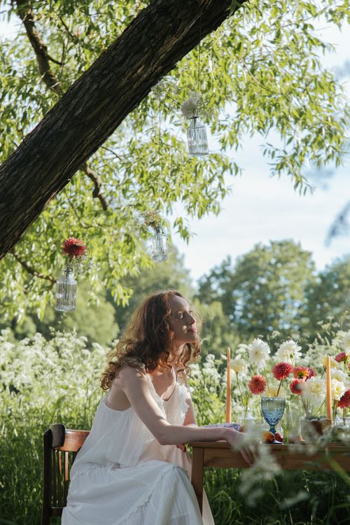 Free A woman in a white dress enjoys a serene picnic in a countryside setting surrounded by flowers. Stock Photo