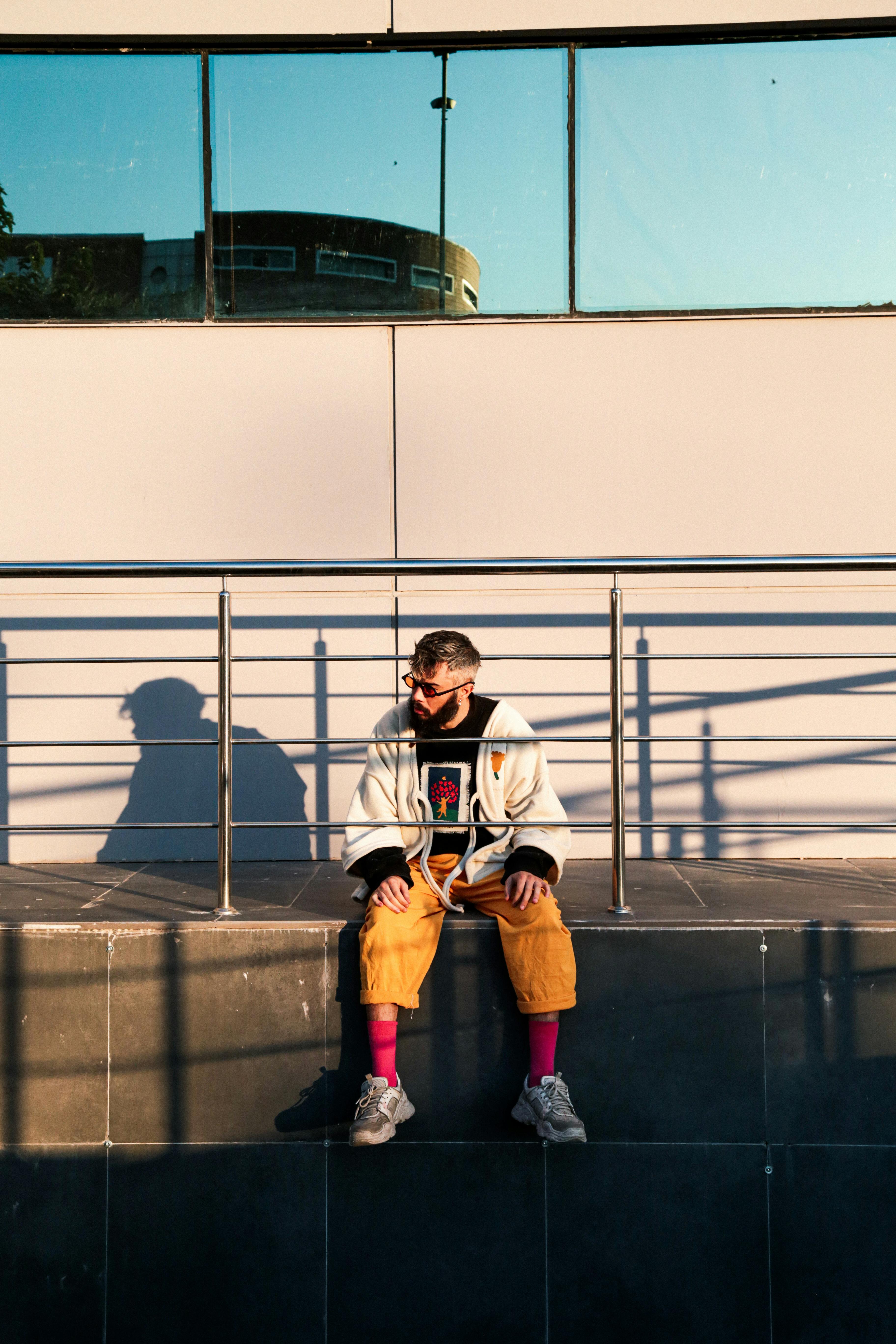 A stylish man sitting next to metal railings on a sunny day, casting shadows.