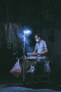 Street vendor cooking at night under a lamp, wearing a mask.