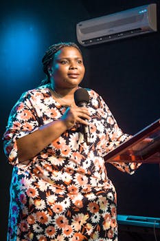Black woman speaking at a podium with a microphone indoors under stage lights.