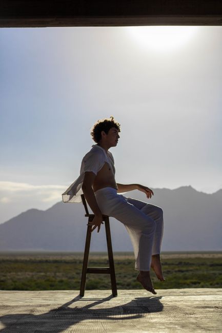 A man in a casual pose on a chair, silhouetted against a mountain view during daytime.