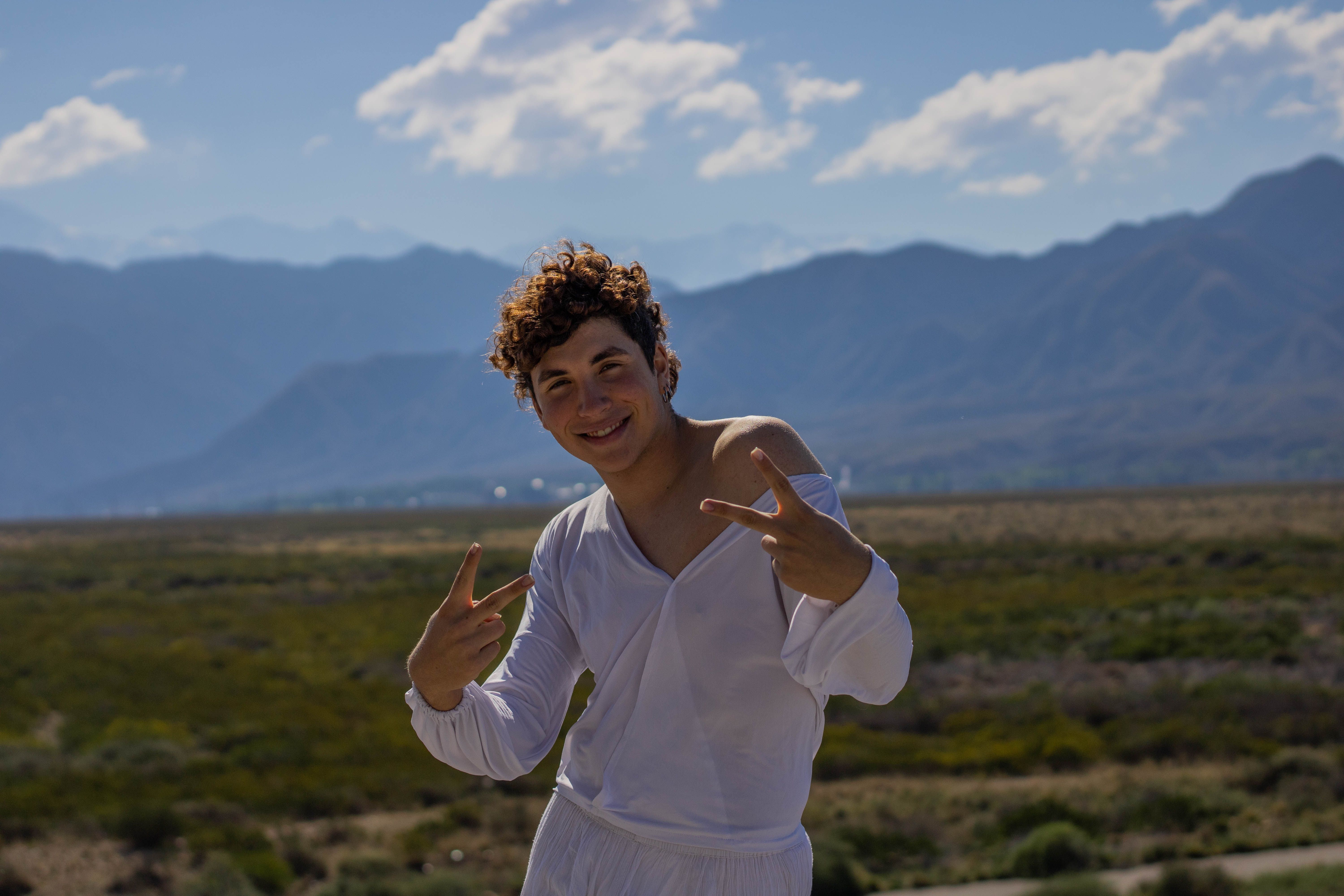 Smiling young man in white clothes, making peace signs outdoors with mountains in the background.