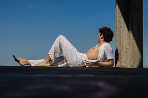 Man in White Shirt and Pants Reclining on Pavement Near Concrete Column