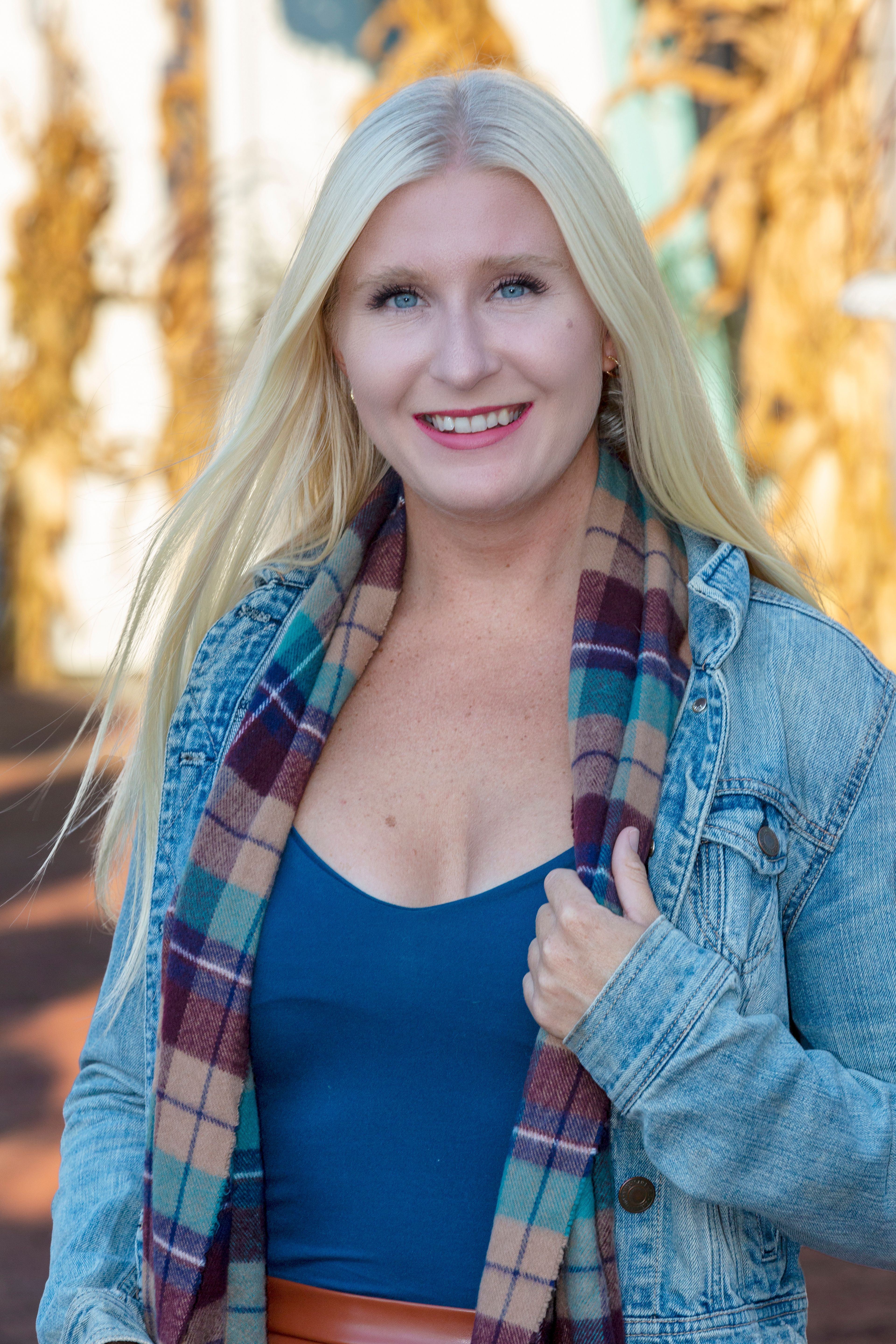 Blonde woman smiling in a denim jacket outdoors with fall background.