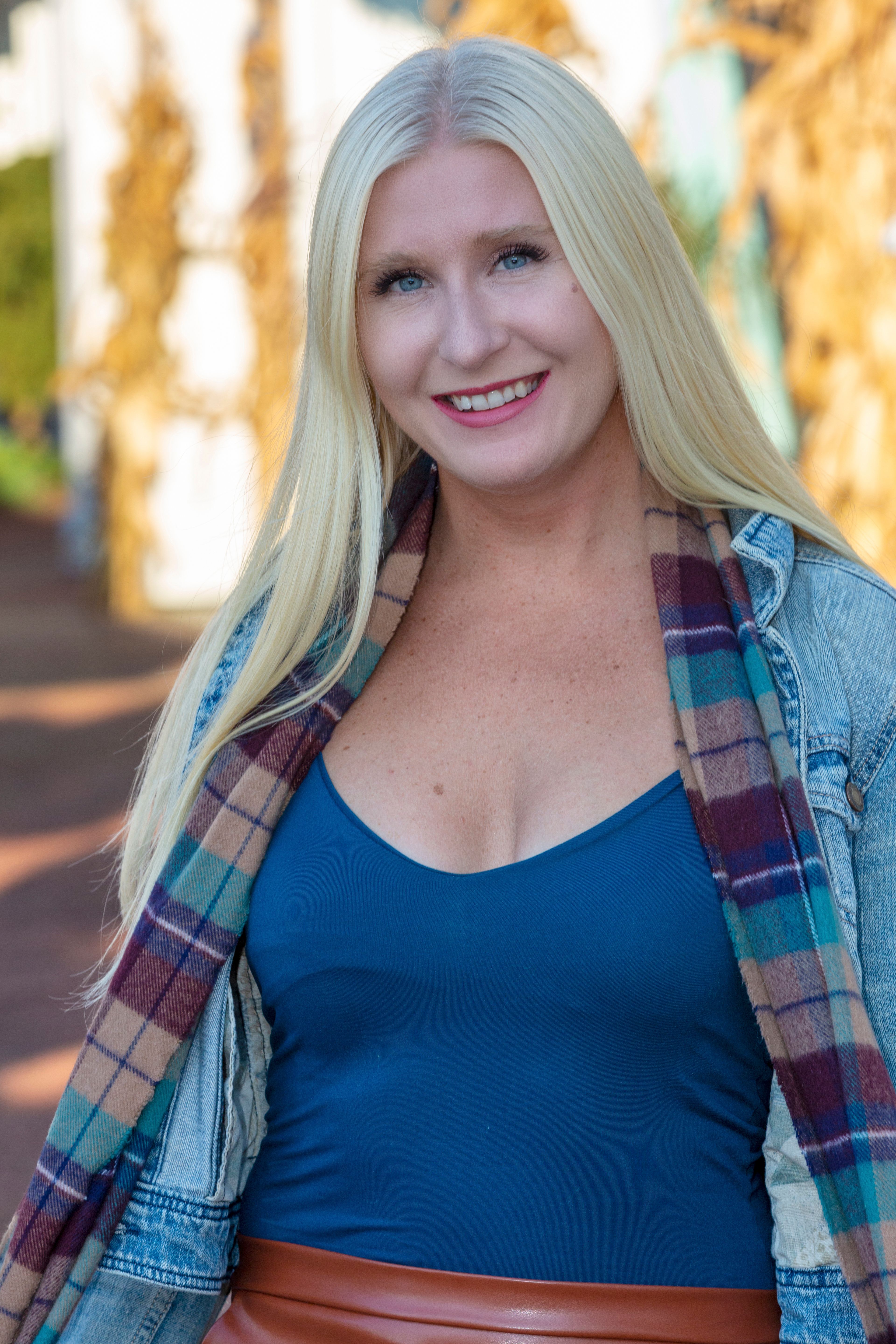Attractive blonde woman smiling outdoors in fall attire, wearing a denim jacket and scarf.