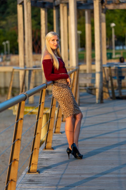 Free A Pretty Woman in Red Off-Shoulder Sleeves and Leopard Print Skirt Leaning on a Metal Railing Stock Photo