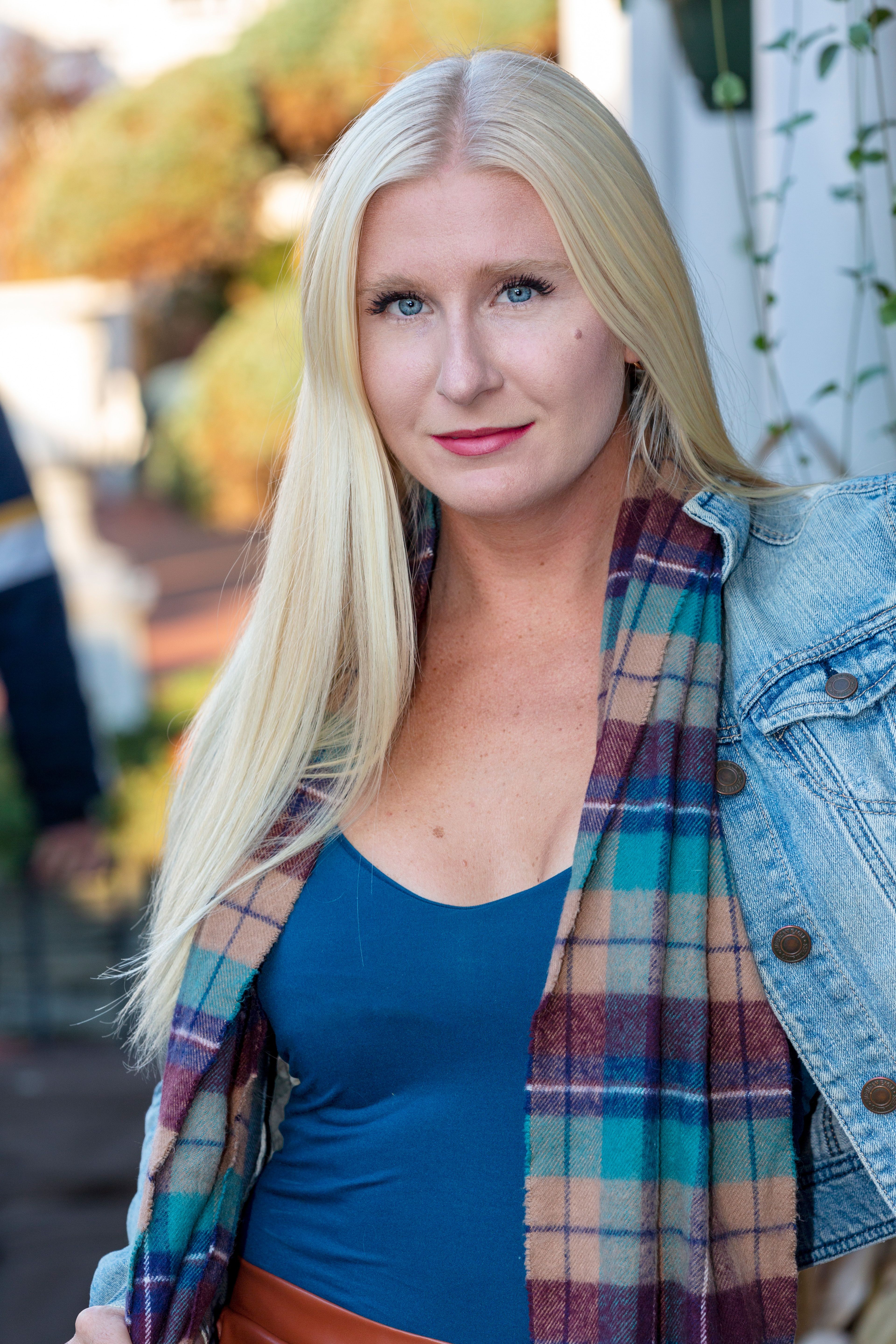 Confident caucasian woman with blonde hair posing outdoors in a denim jacket and plaid scarf.