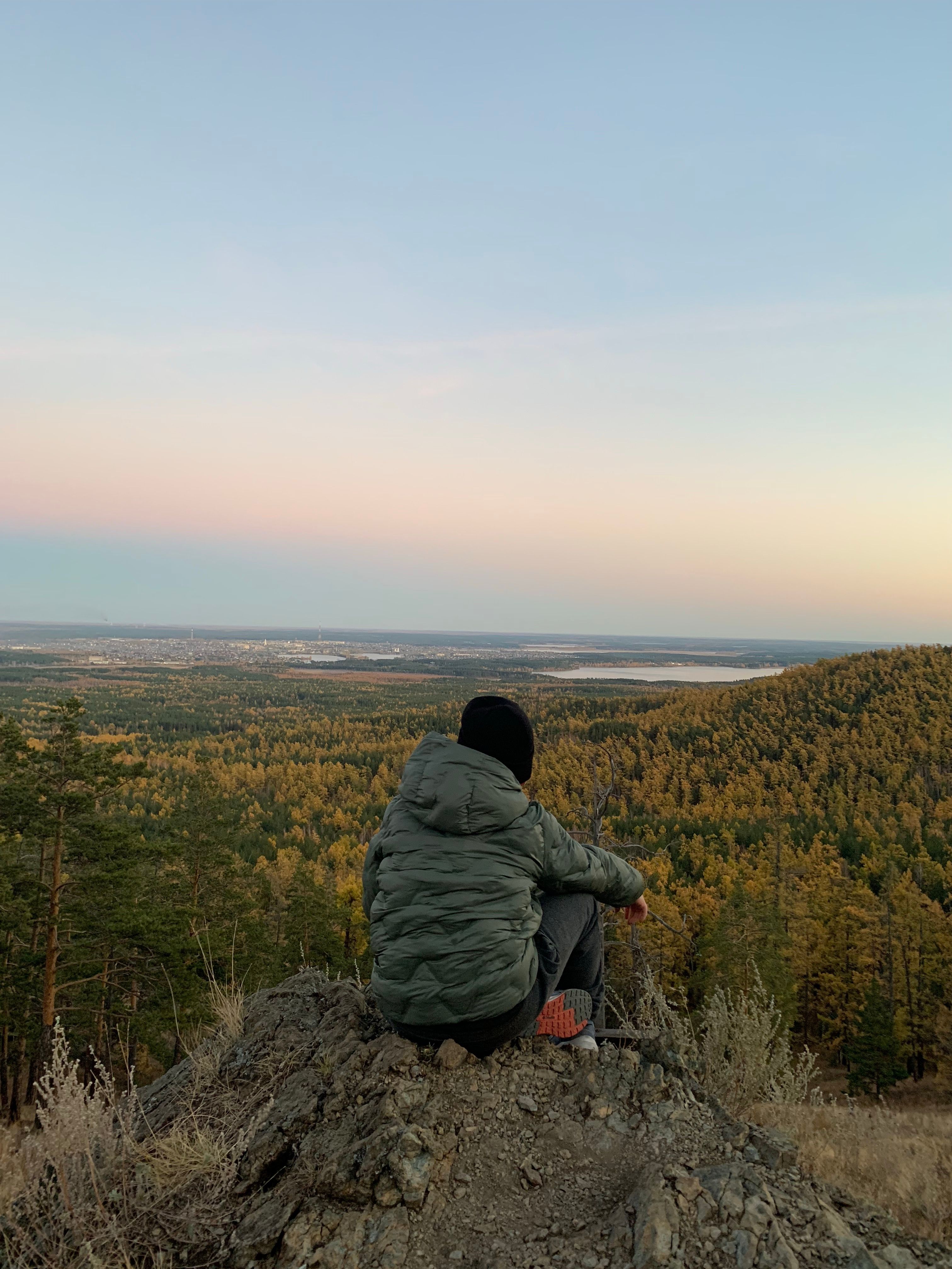 A person sits thoughtfully on a rocky hill overlooking a vast scenic valley at sunset.