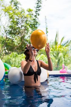 Woman in bikini plays with balloons in a pool, wearing sunglasses on a sunny day.
