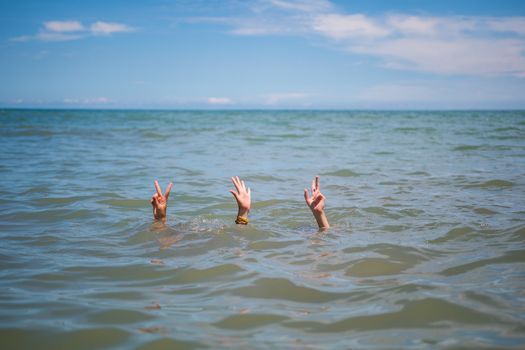 Two hands showing peace signs rise above the ocean water under a clear blue sky.