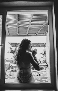 Black and white photo of a woman in a bikini enjoying a drink by the poolside.