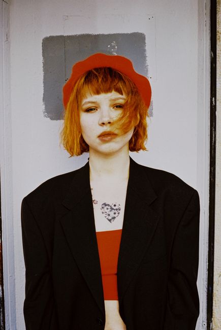 Portrait of a woman with red hair and a tattoo wearing a black blazer and red hat.