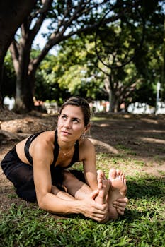 Fit woman doing yoga stretches on grass in a sunny park with trees around her.
