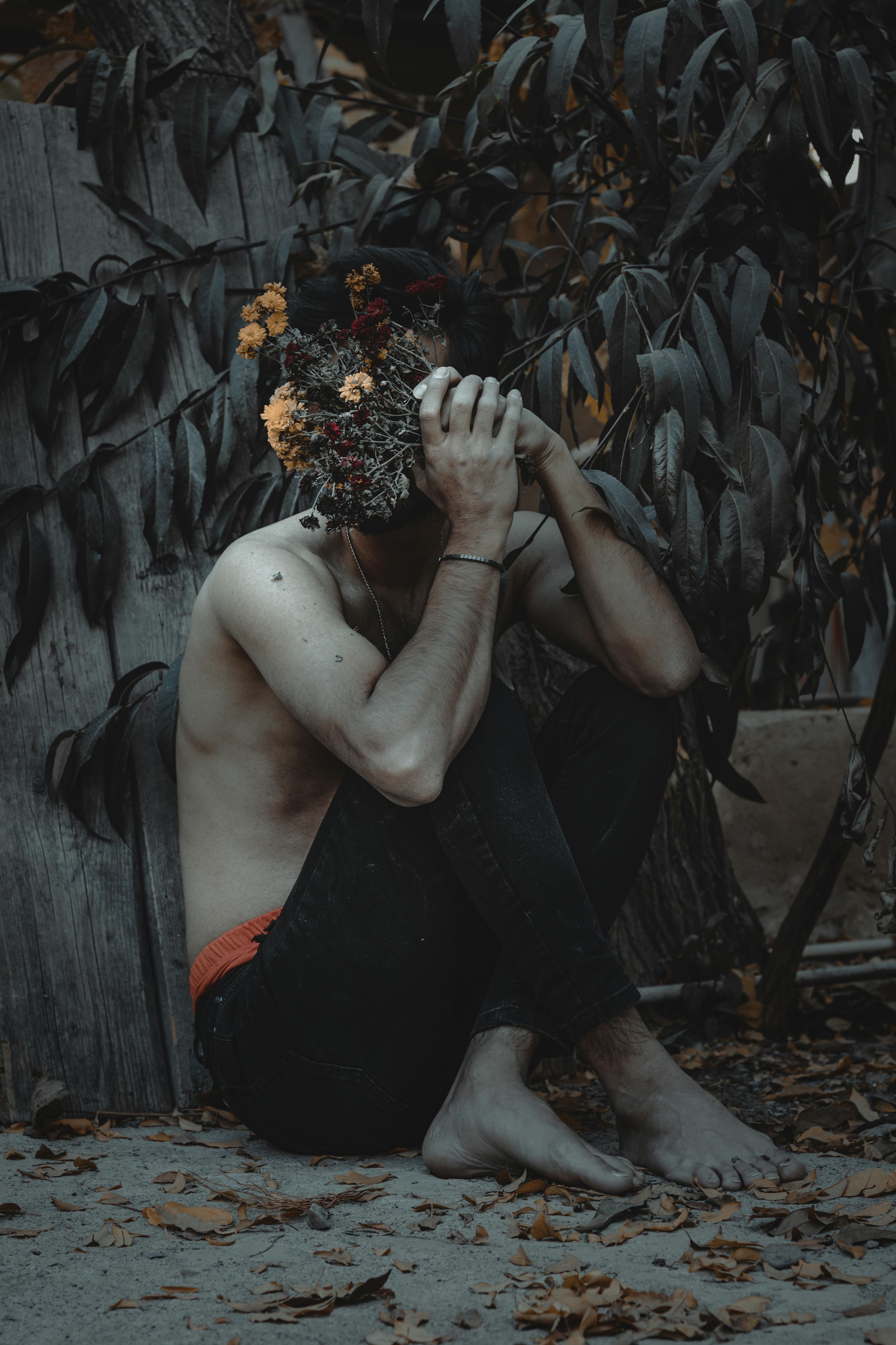 Shirtless man sitting barefoot outdoors with a flower-covered face amidst autumn leaves.
