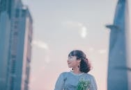 画中一位微笑的妇女手持鲜花，站在户外，背景是城市的摩天大楼