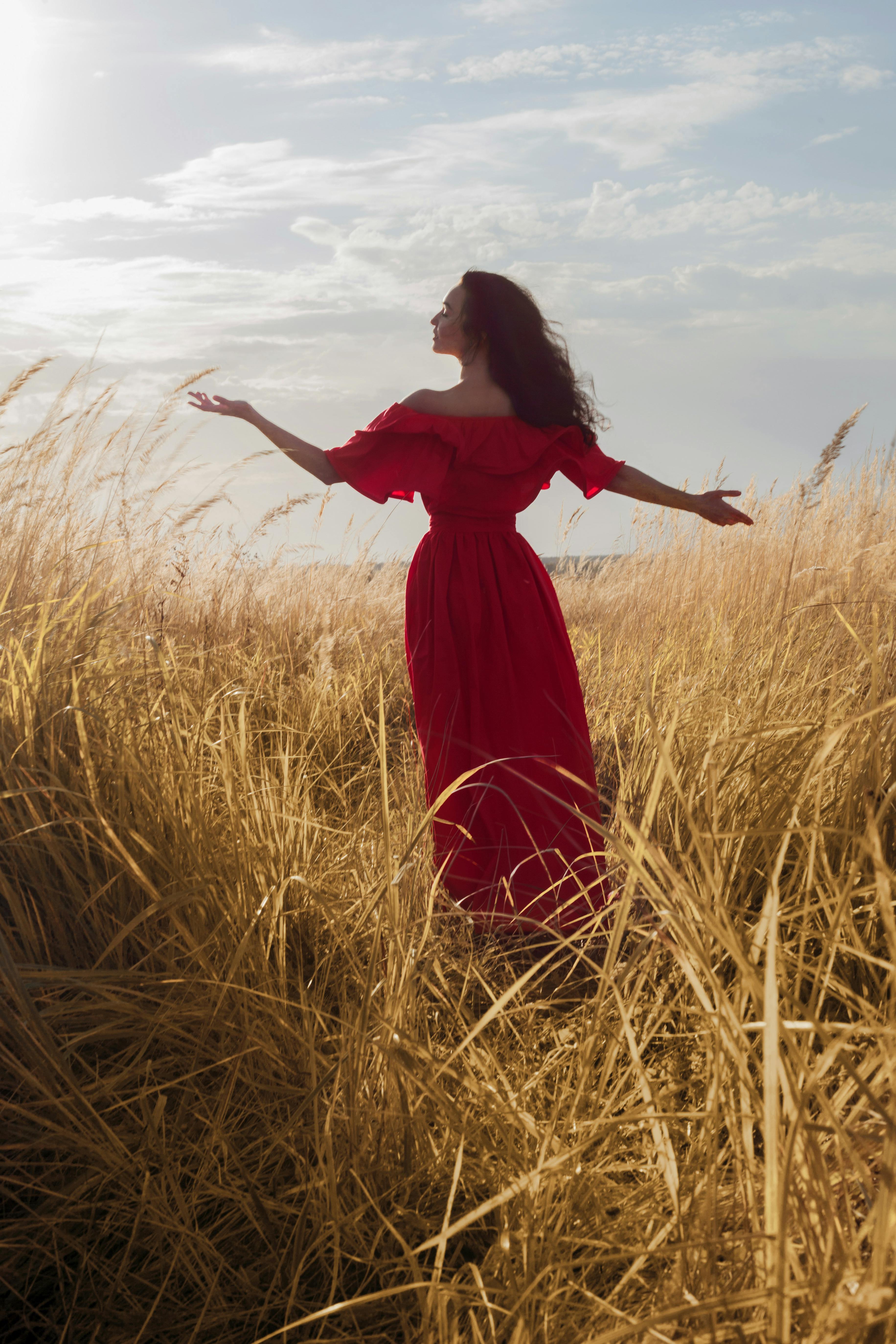 attractive woman in long red dress standing in tall grass and greeting sunlight