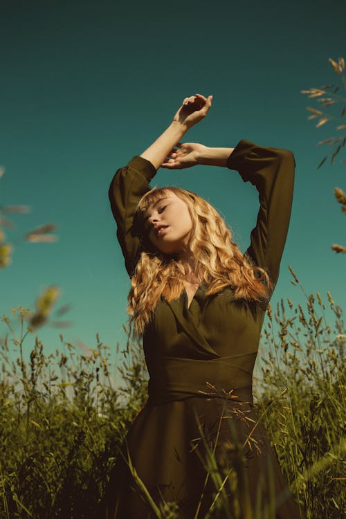 Free Blond Woman in Dress Standing in Green Meadow with Arms Raised in Ecstasy Stock Photo