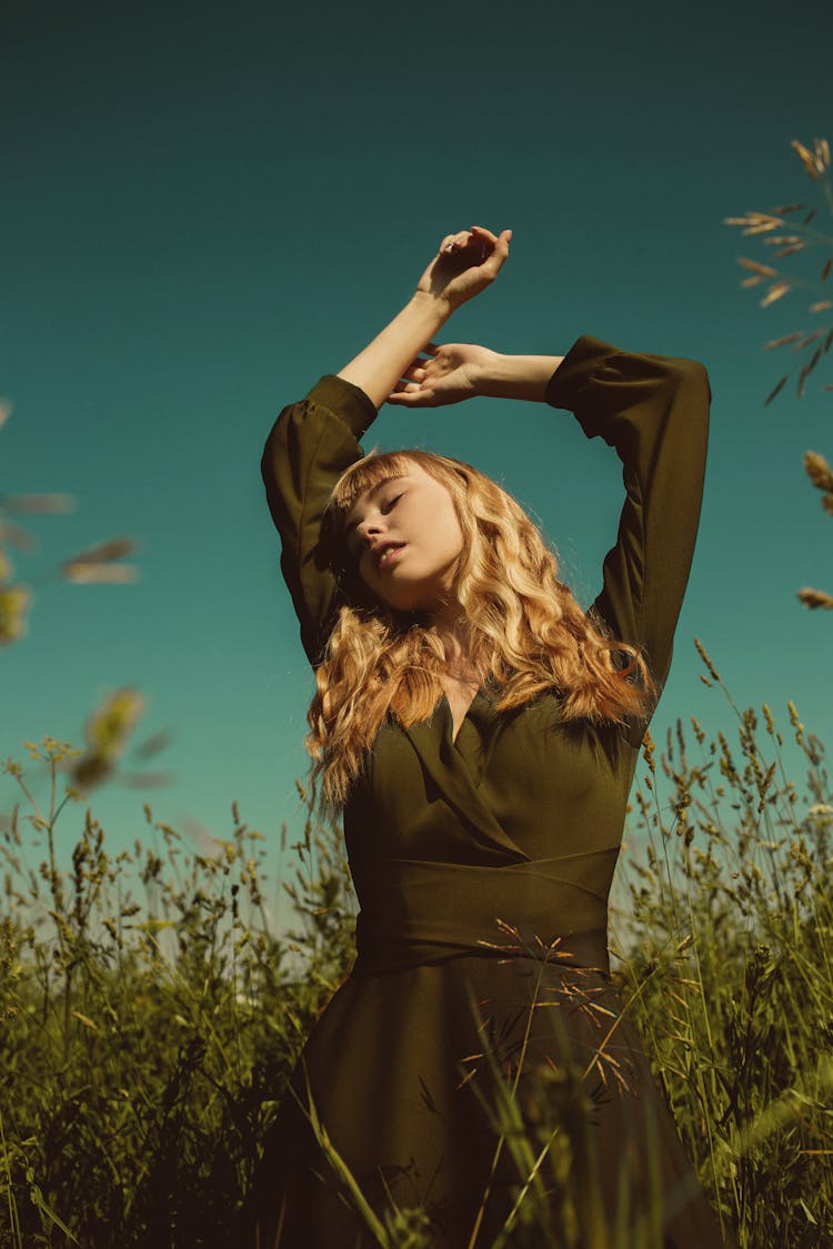 Blond Woman In Dress Standing In Green Meadow With Arms Raised In Ecstasy