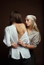 Blond Woman in Kerchief Embracing Brunette Woman and Fastening Button of Her Shirt