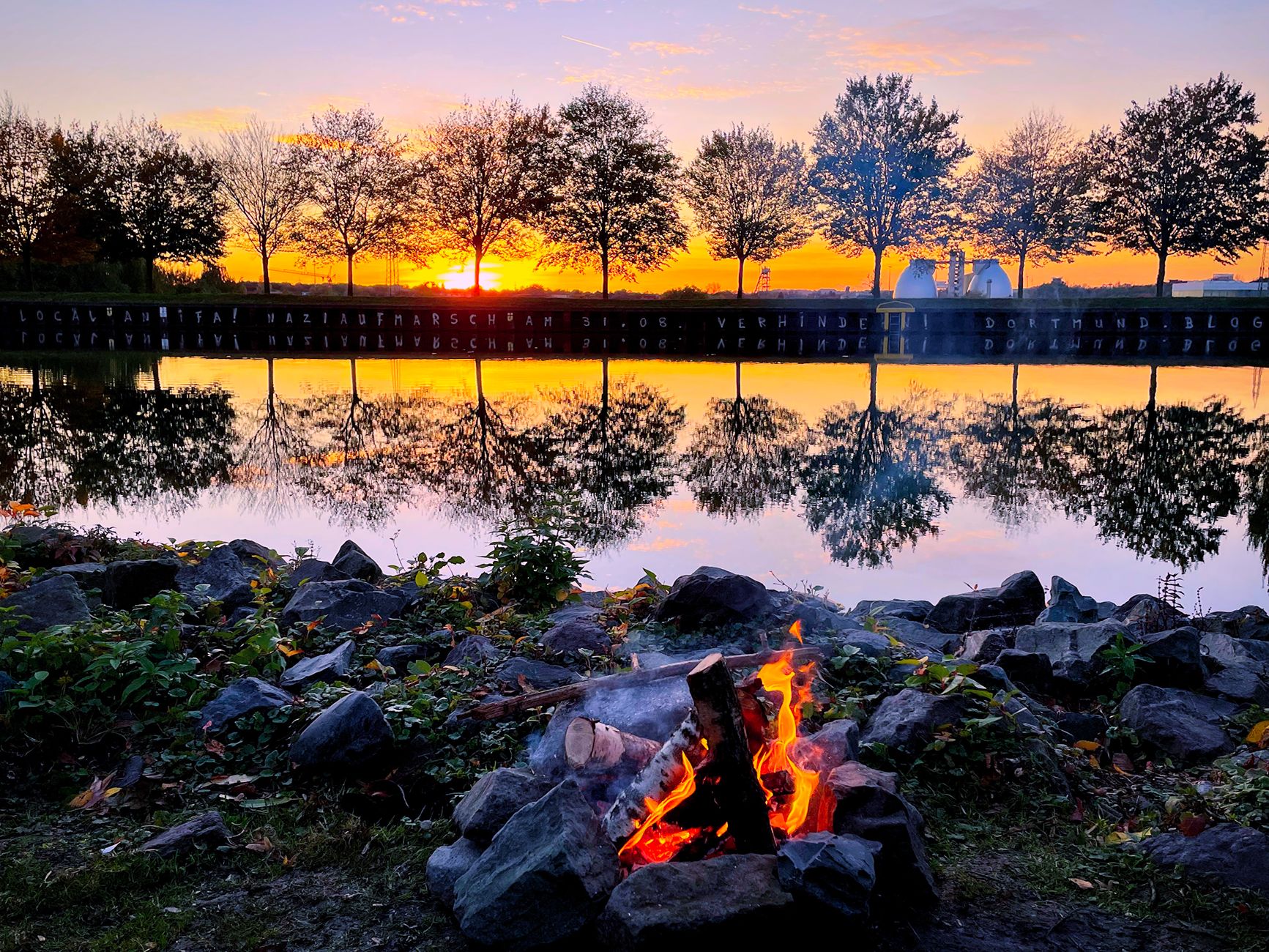 Campfire burning beside a quiet lake at sunset