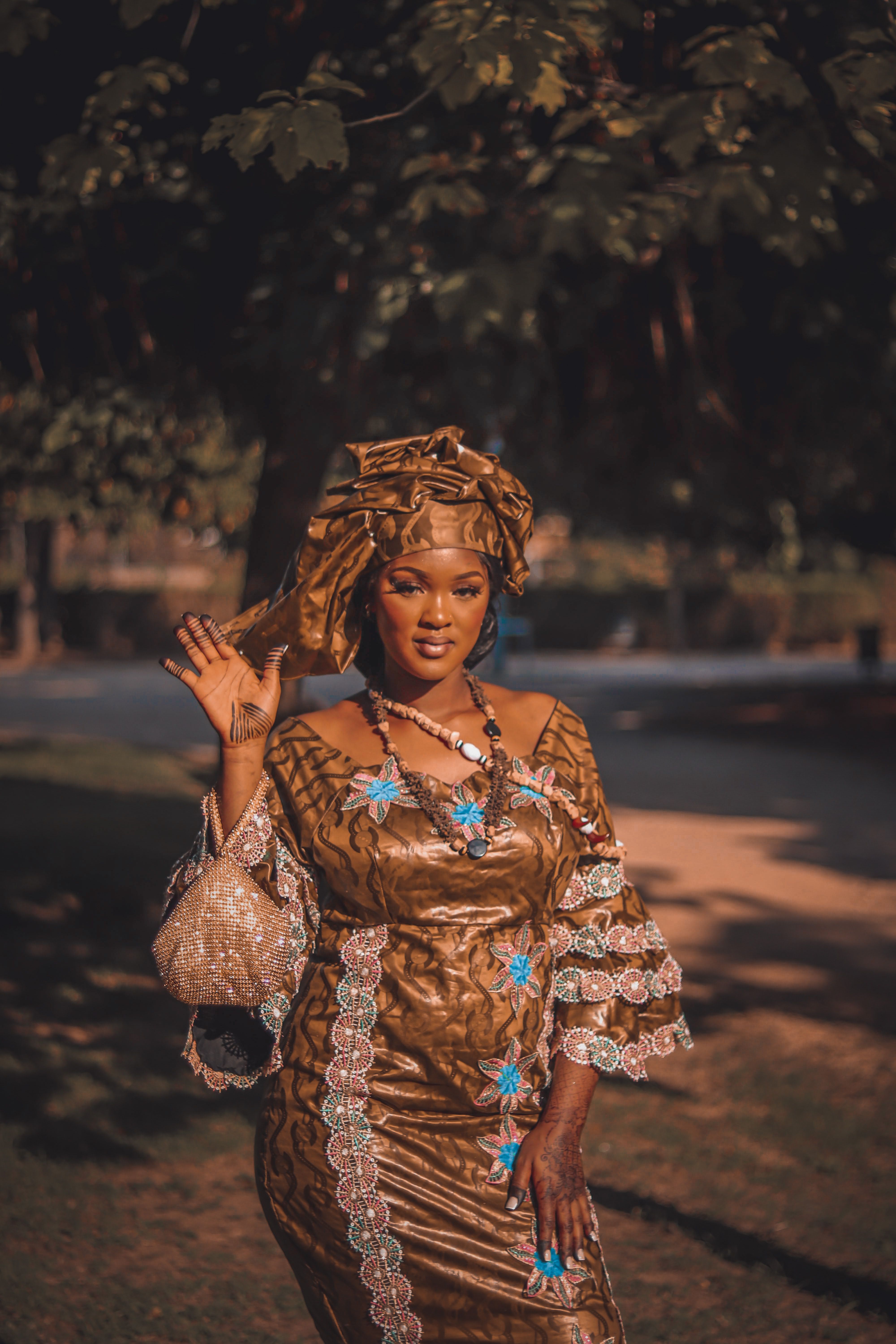 A portrait of an African woman in a traditional brown dress waving outdoors with trees in the background.