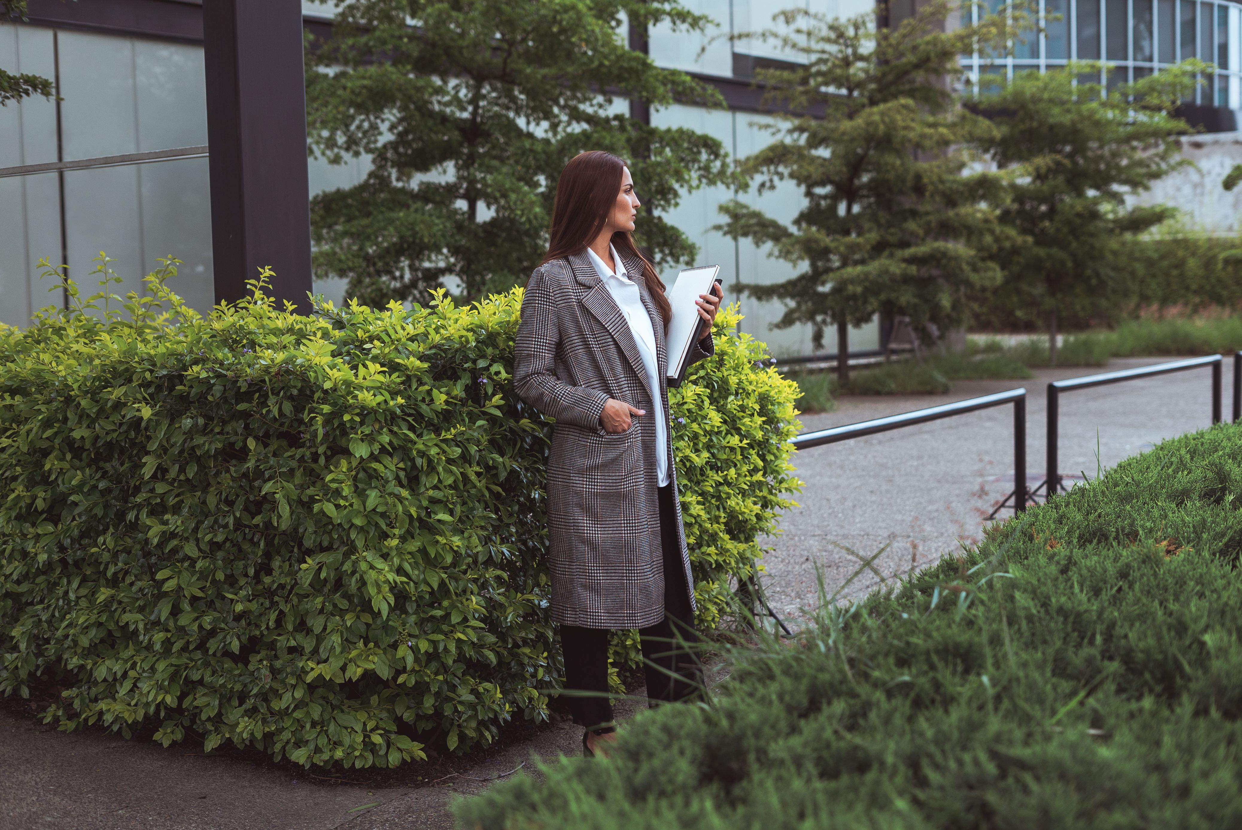 A woman in a coat stands outdoors with documents, surrounded by greenery.