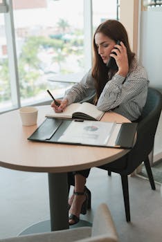 Woman in striped shirt multitasking in an office, writing notes while on a phone call, business setting.
