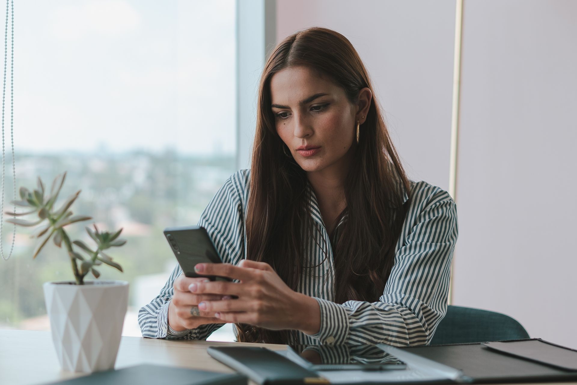 A businesswoman attentively using her smartphone at a desk in a modern office setting with a potted plant.