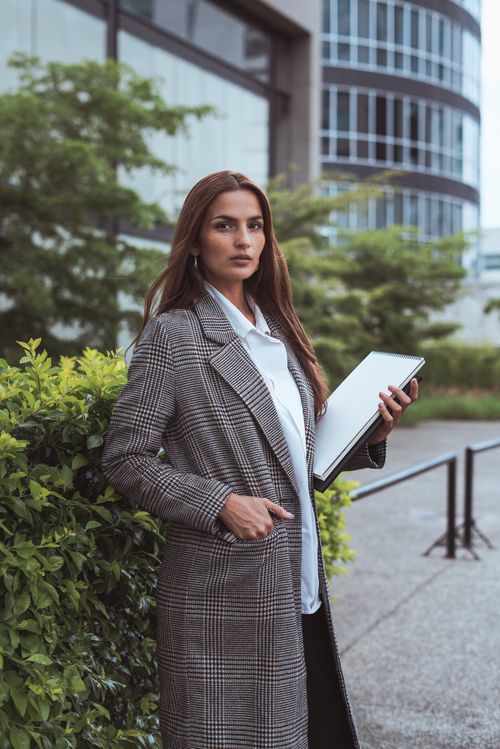 Free Confident woman in a stylish coat standing outside an office building holding a laptop. Stock Photo