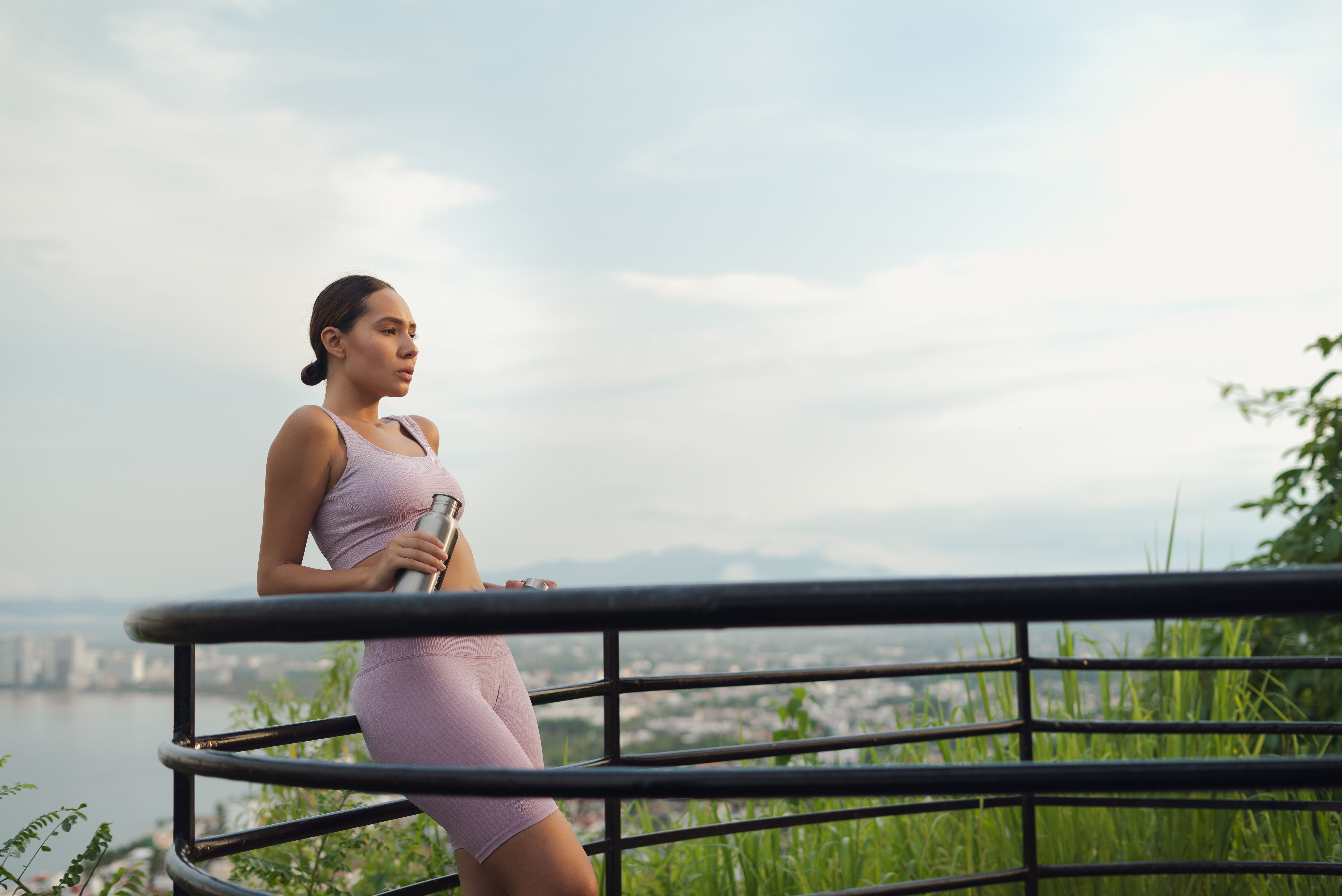 Woman in athletic wear holding a thermos by a railing overlooking a cityscape.