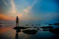 一名女子站在岩石上，望着宁静湖面上那轮充满活力的日落，倒影完美无瑕