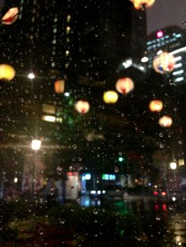 A vibrant city scene on a rainy night with raindrops on the window and glowing lanterns.