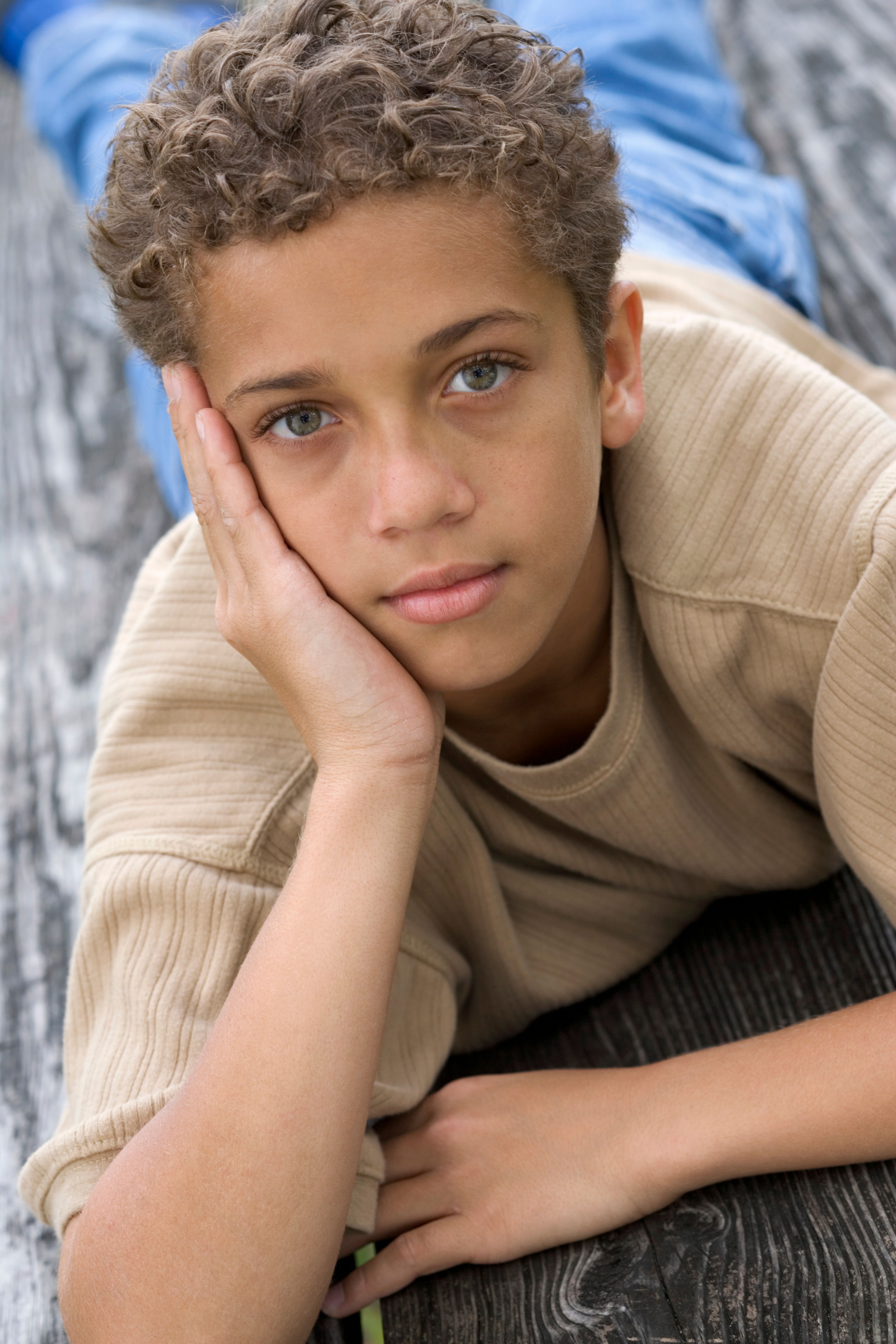 A young boy with curly hair rests on a wooden floor, looking thoughtful and relaxed.