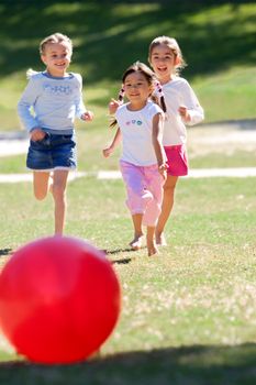 Three young girls joyfully chase a large red ball outdoors on a sunny day.