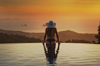 在泰国的无边泳池里，一名戴着太阳帽的女子正欣赏着宁静的日落景色