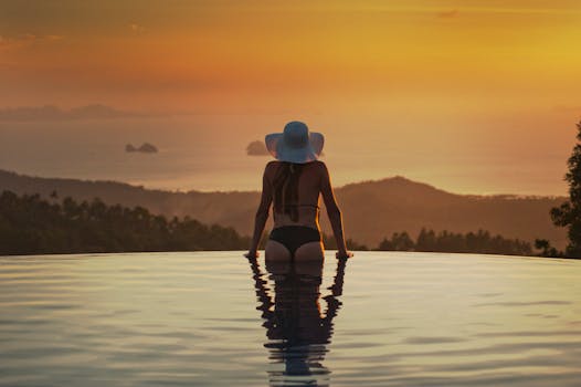 在泰国的无边泳池里，一名戴着太阳帽的女子正欣赏着宁静的日落景色