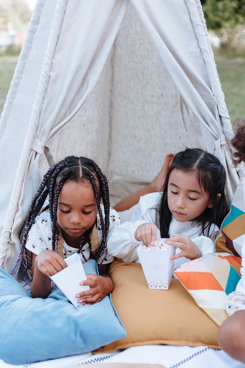 Free Two young girls relaxing and sharing popcorn inside a cozy teepee tent outdoors. Stock Photo