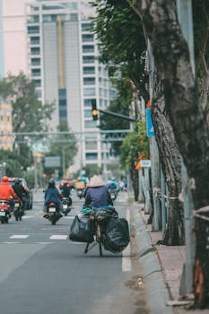 A cyclist transporting bags on a city street amid urban traffic and tall buildings.