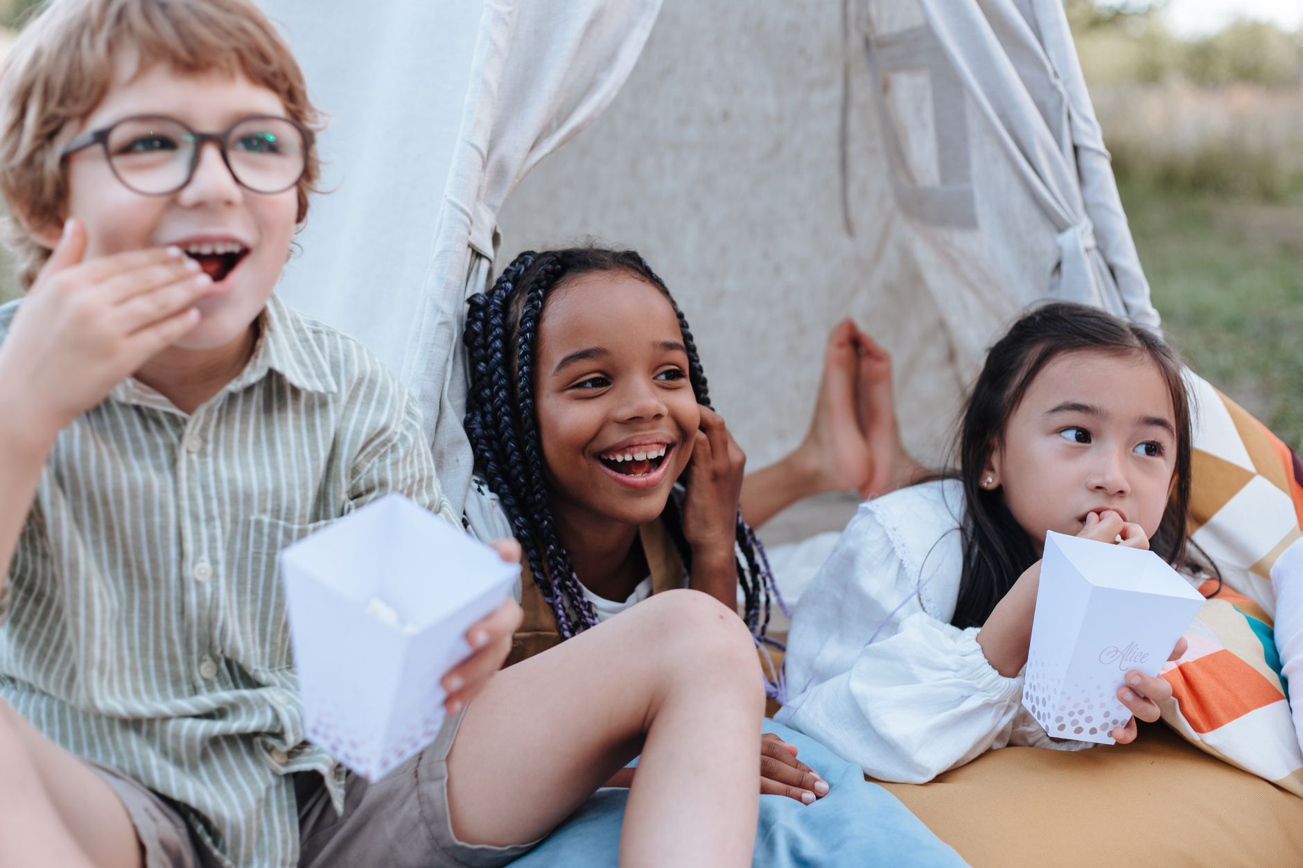 Three diverse children enjoying popcorn in a backyard tent. Perfect summer leisure activity.