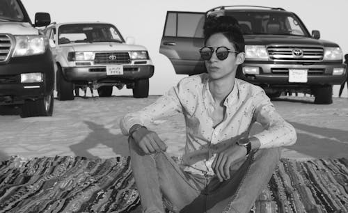 Man Sitting on a Blanket in a Desert with SUVs Parked in the Background 