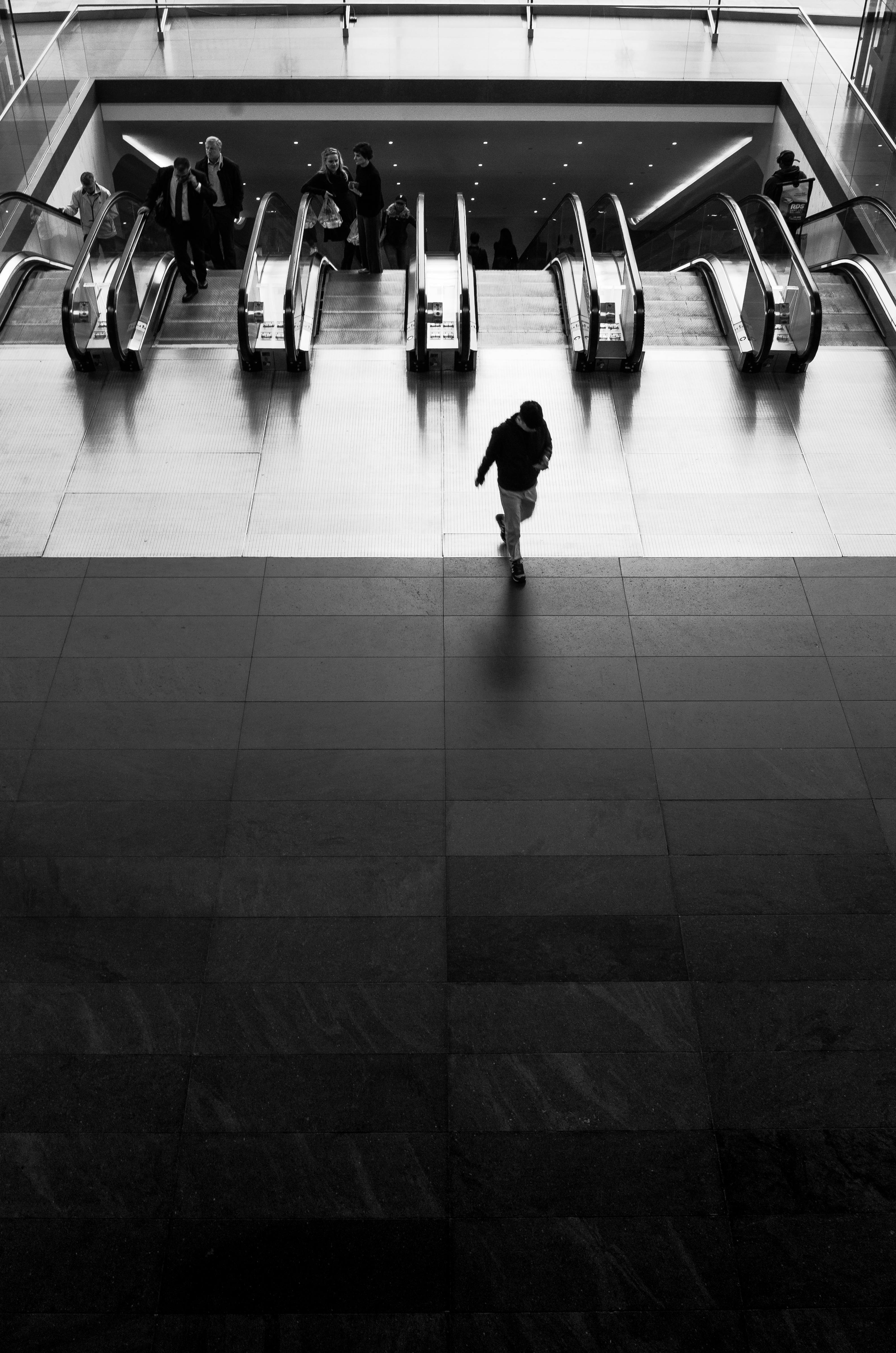 Monochrome image of a lone person approaching escalators in busy urban setting.