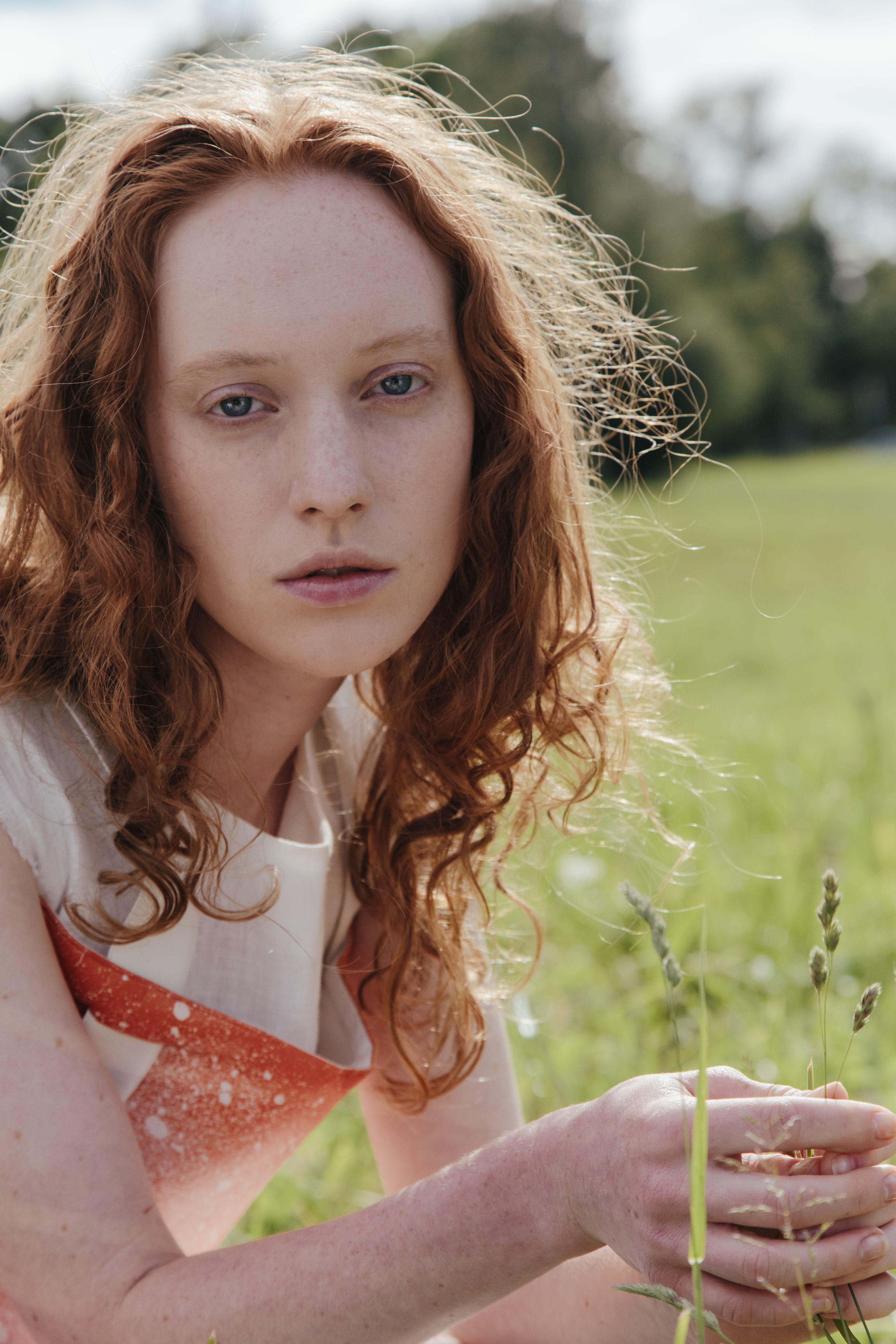 A woman with red hair poses thoughtfully in a sunny green field.