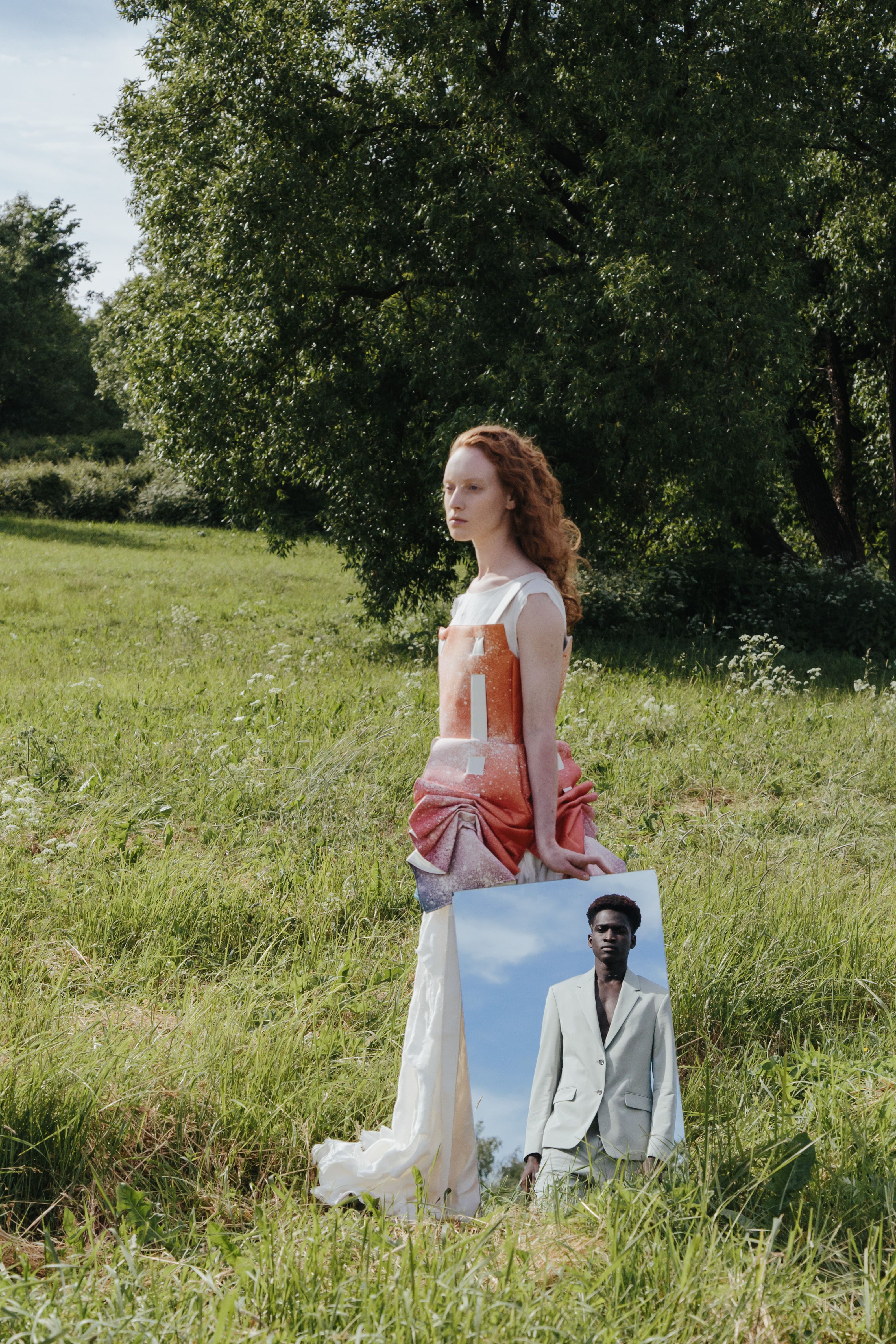 A woman in a field holding a mirror reflecting a man in a suit, showcasing nature and fashion elements.